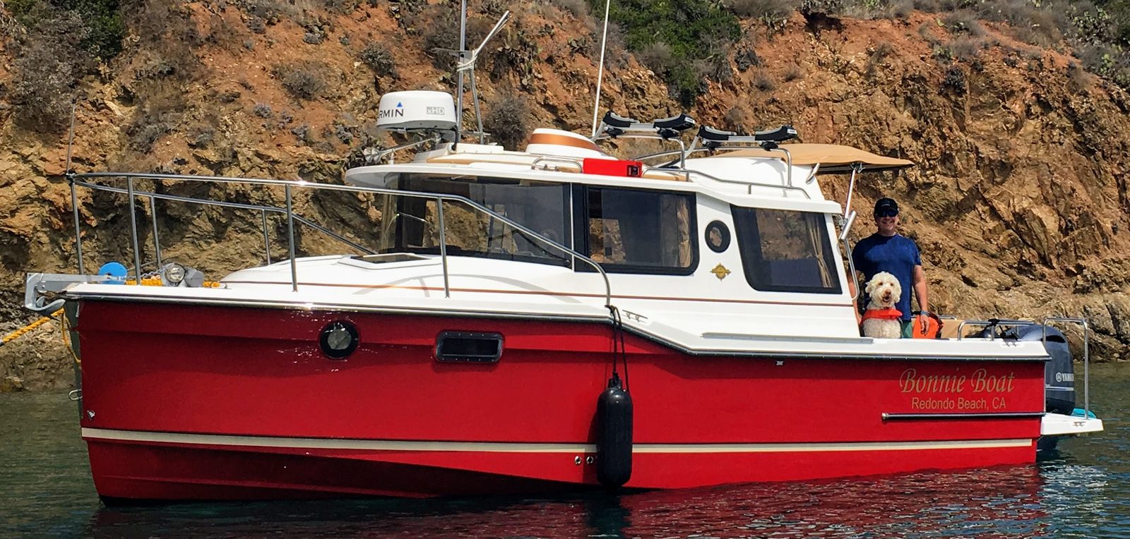 A red trawler-style motorboat at anchor in a rocky cove, a person and a small white dog in the cockpit
